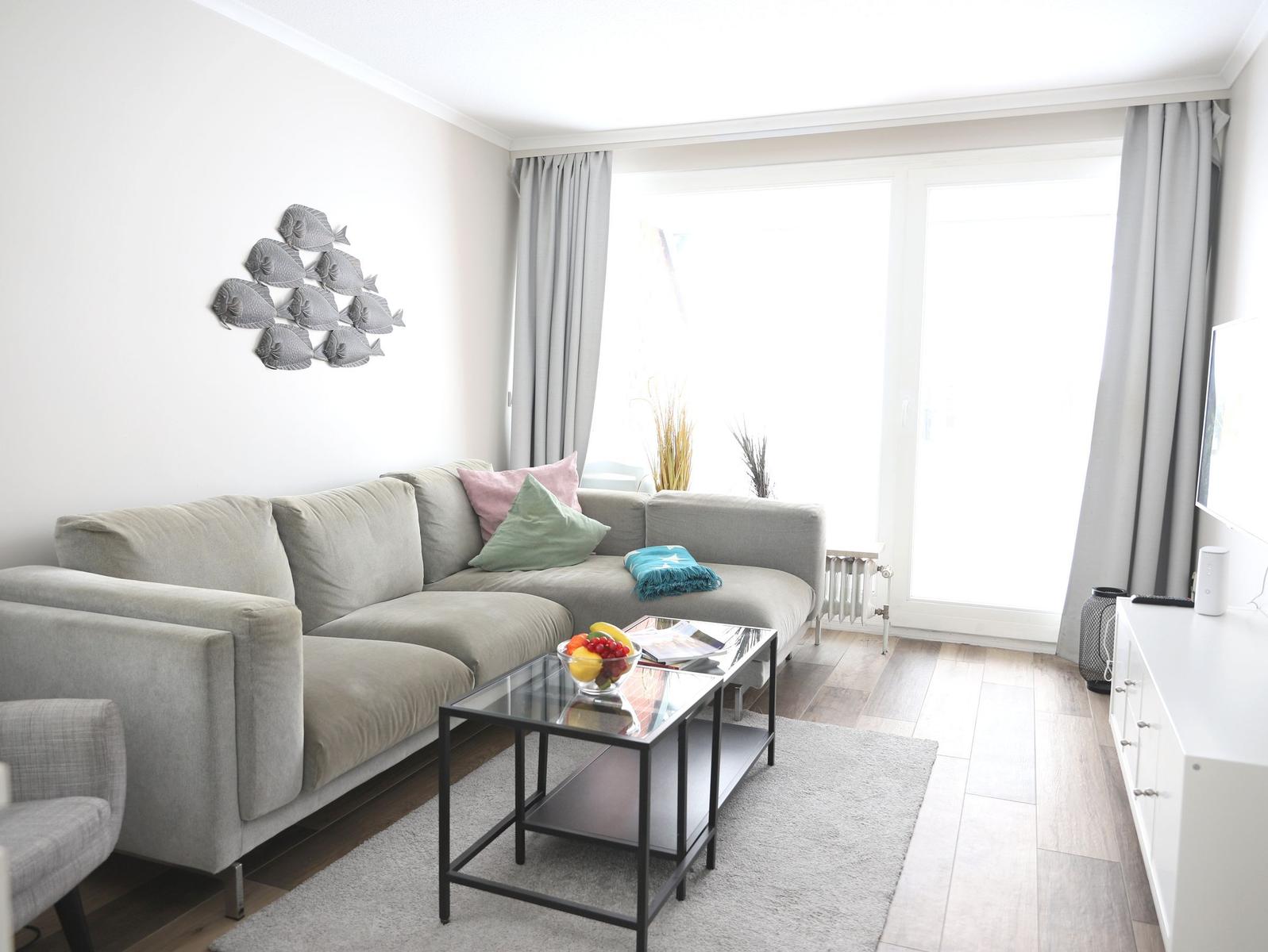 Wohnzimmer mit grauem Sofa, Glas-Tisch und großen Fenstern.