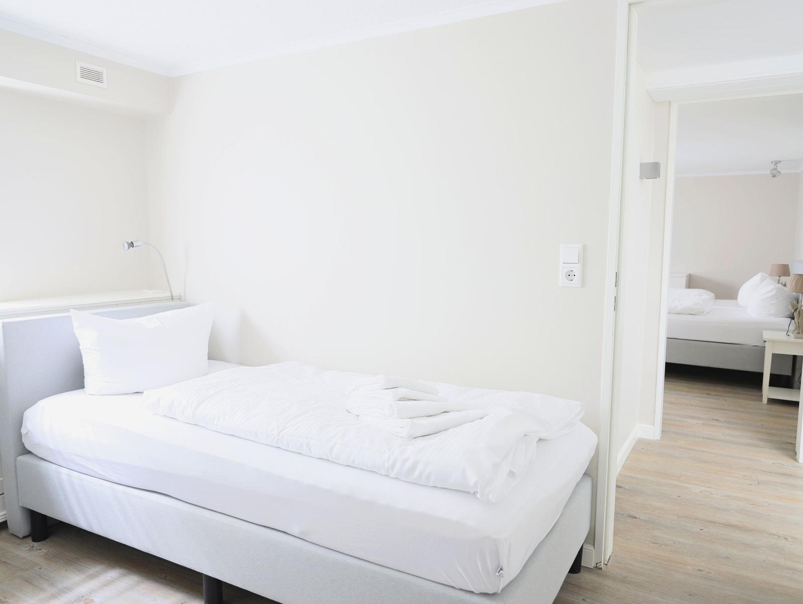 Ein Schlafzimmer mit weißem Bett und Holzboden. Durchgang zu einem zweiten Schlafzimmer.