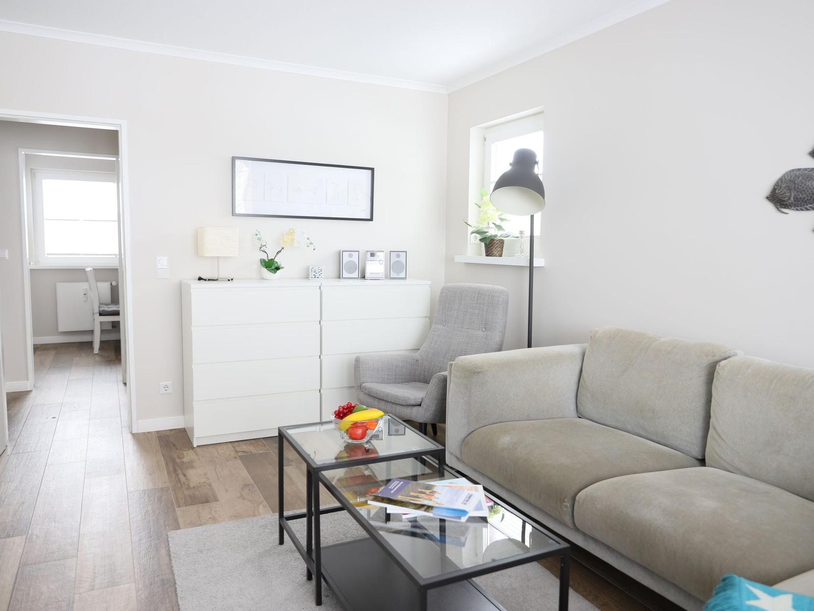 Wohnzimmer mit Sofa, Sessel, Glas-Tisch und weißen Schränken. Durchgang zum Schlafzimmer.