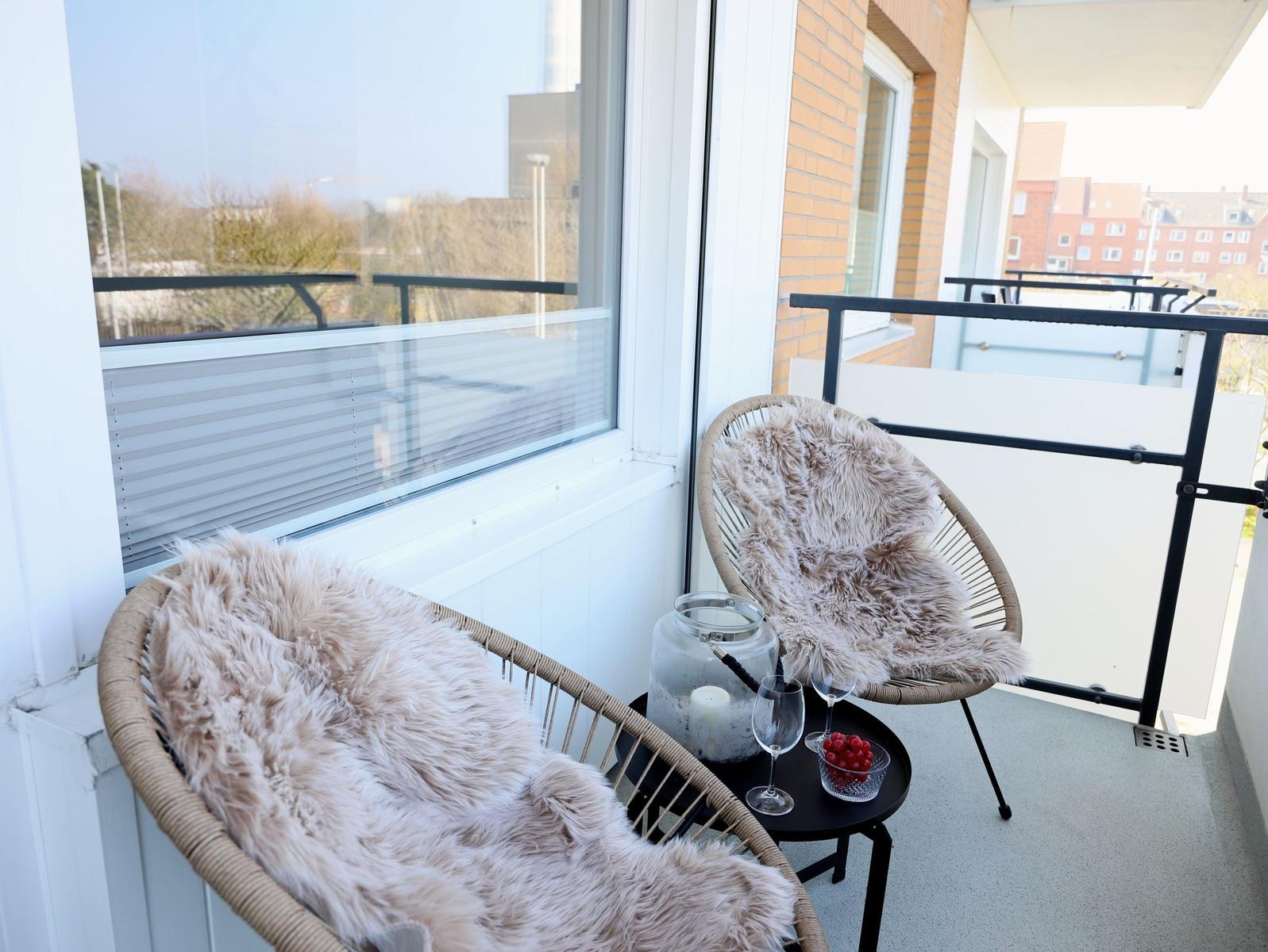 Balcony with two wicker chairs, fur throws, small table, and glasses. View of buildings and trees.