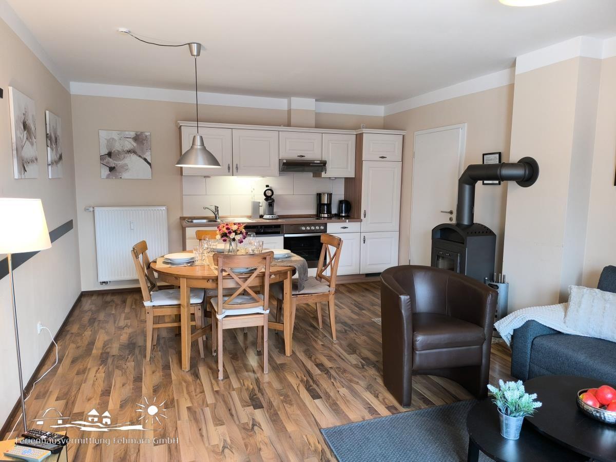 Ein Essbereich mit Holztisch und Stühlen, Küche mit weißen Schränken und Wohnbereich mit Sofa und Sessel.