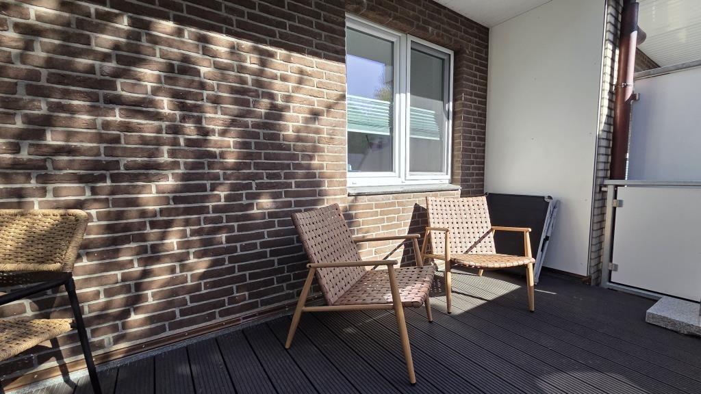 Terrace with two chairs, wooden decking, and brick wall. Window and railing are visible.