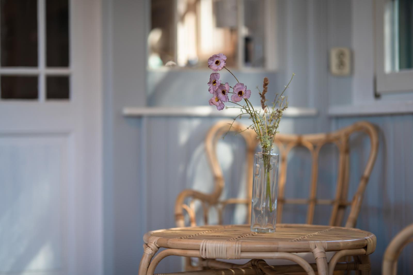 Rattan-Tisch mit Glasvase und Blumen vor blauen Wänden und Stühlen.