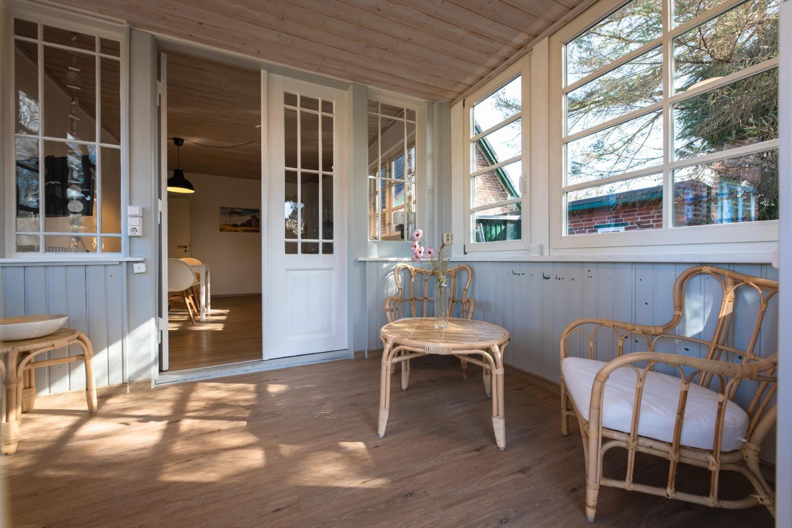Terrasse mit Rattanmöbeln, Holzboden und Fenstern. Tür führt ins Innere.