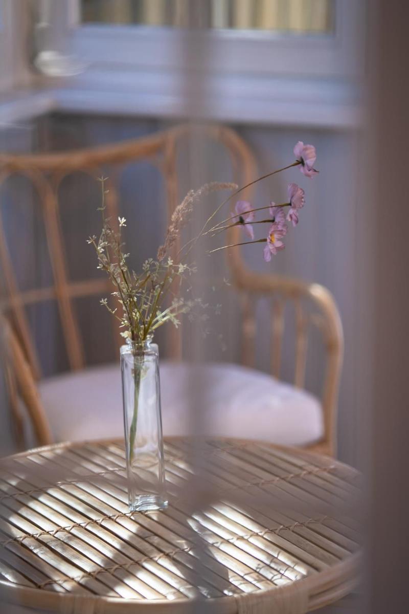 Auf dem Tisch steht eine Vase mit Blumen. Im Hintergrund sind Stühle und ein Fenster zu sehen.