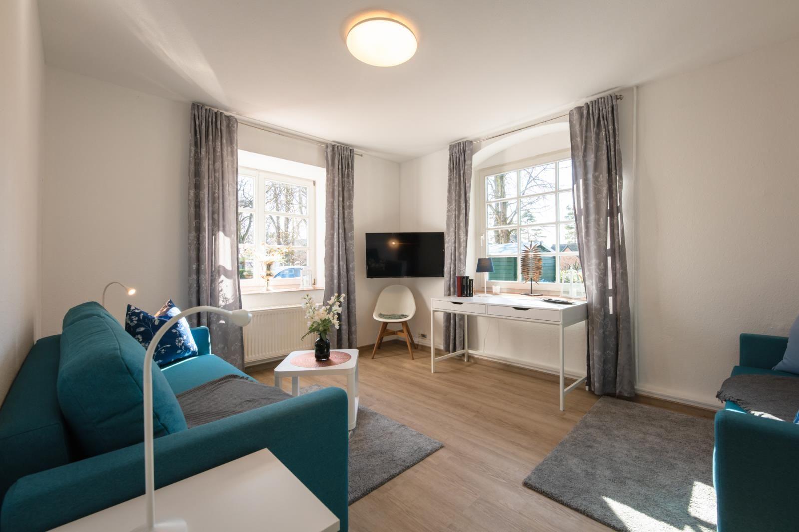 Wohnzimmer mit Sofa, Schreibtisch und TV. Zwei Fenster mit Vorhängen.
