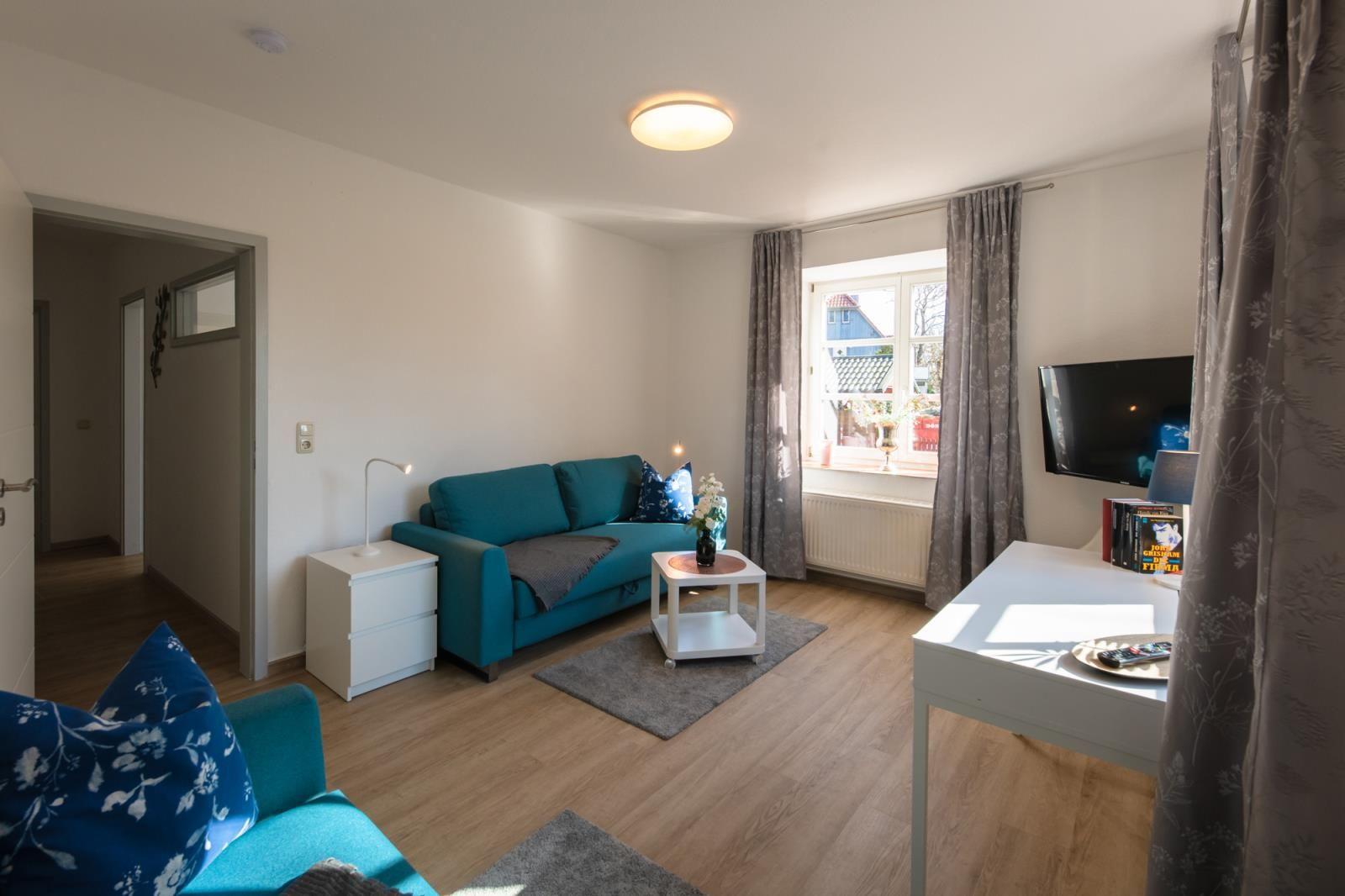 Wohnzimmer mit Sofa, Schreibtisch und Fernseher. Fenster mit Vorhängen. Holzboden.