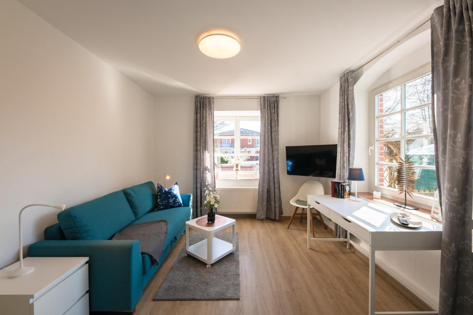 Ein Wohnzimmer mit Sofa, Schreibtisch und Fernseher. Holzboden und große Fenster.