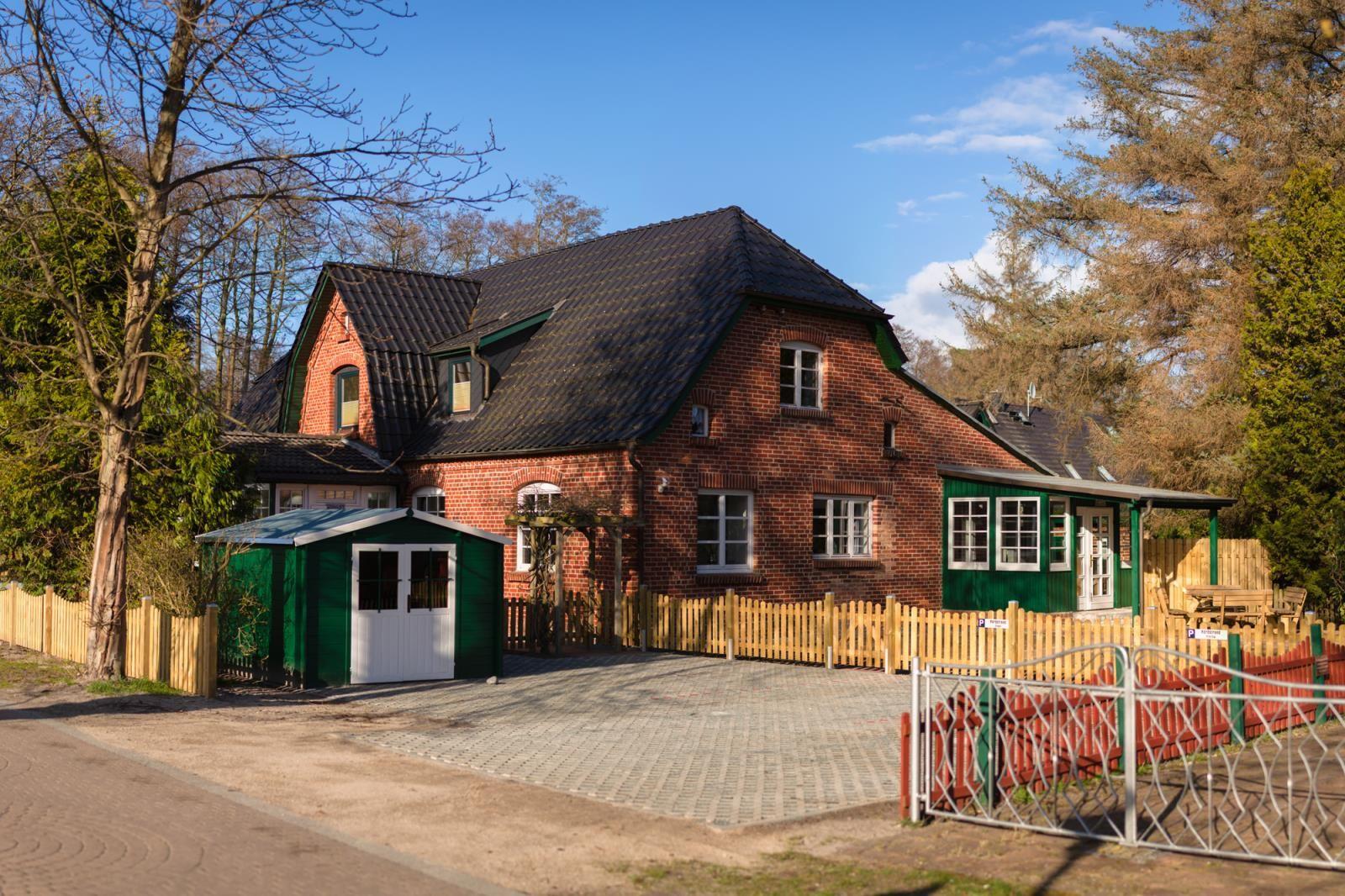 Rotes Backsteinhaus mit grünem Anbau und gelbem Zaun. Vorgarten mit Pavillon und Parkplatz.