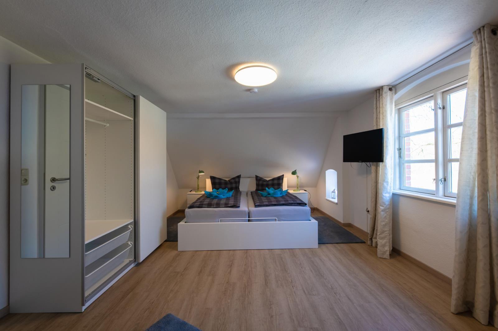 Bedroom with double bed, wardrobe, and TV. Wooden floor and window with curtains.