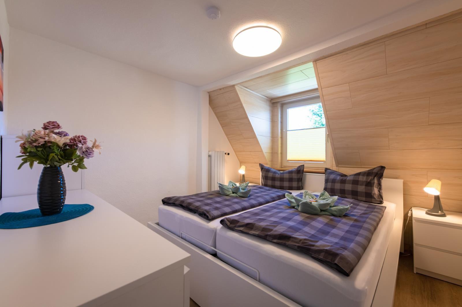 Schlafzimmer mit Doppelbett, Holzwand und Fenster. Auf Tisch Blumen in Vase.