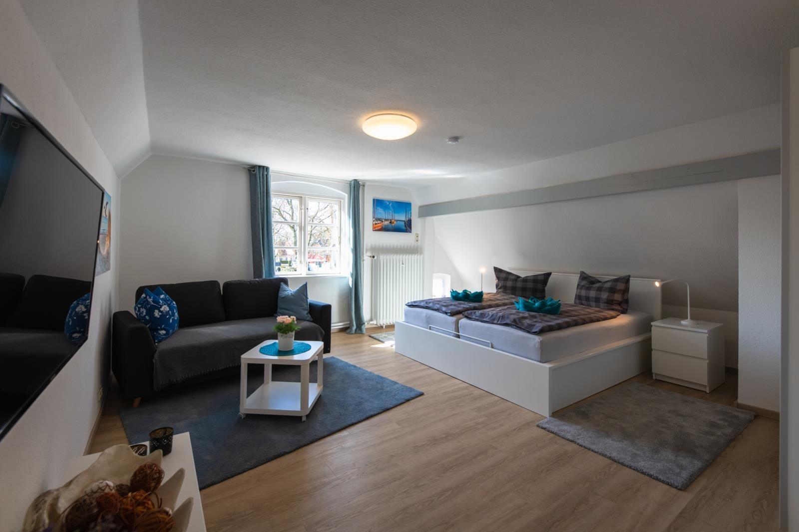 Schlafzimmer mit Doppelbett, Sofa und Fenster. Holzboden und weiße Wände.
