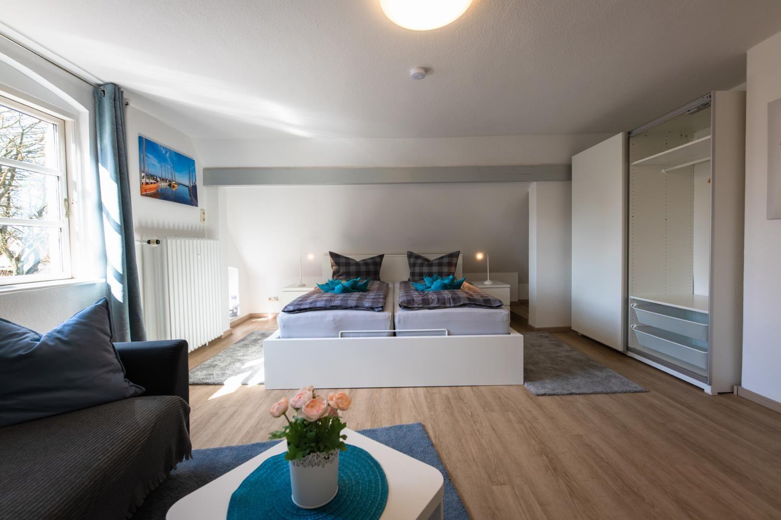 Zimmer mit Doppelbett, Schrank und Fenster. Tisch mit Blumen im Vordergrund.