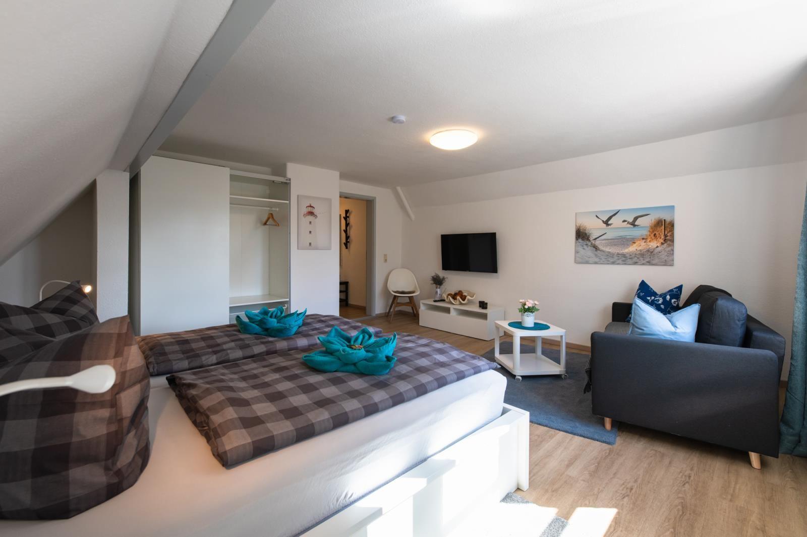 Schlafzimmer mit Doppelbett, Sofa und Fernseher. Ankleidezimmer und Wandbilder sind sichtbar.