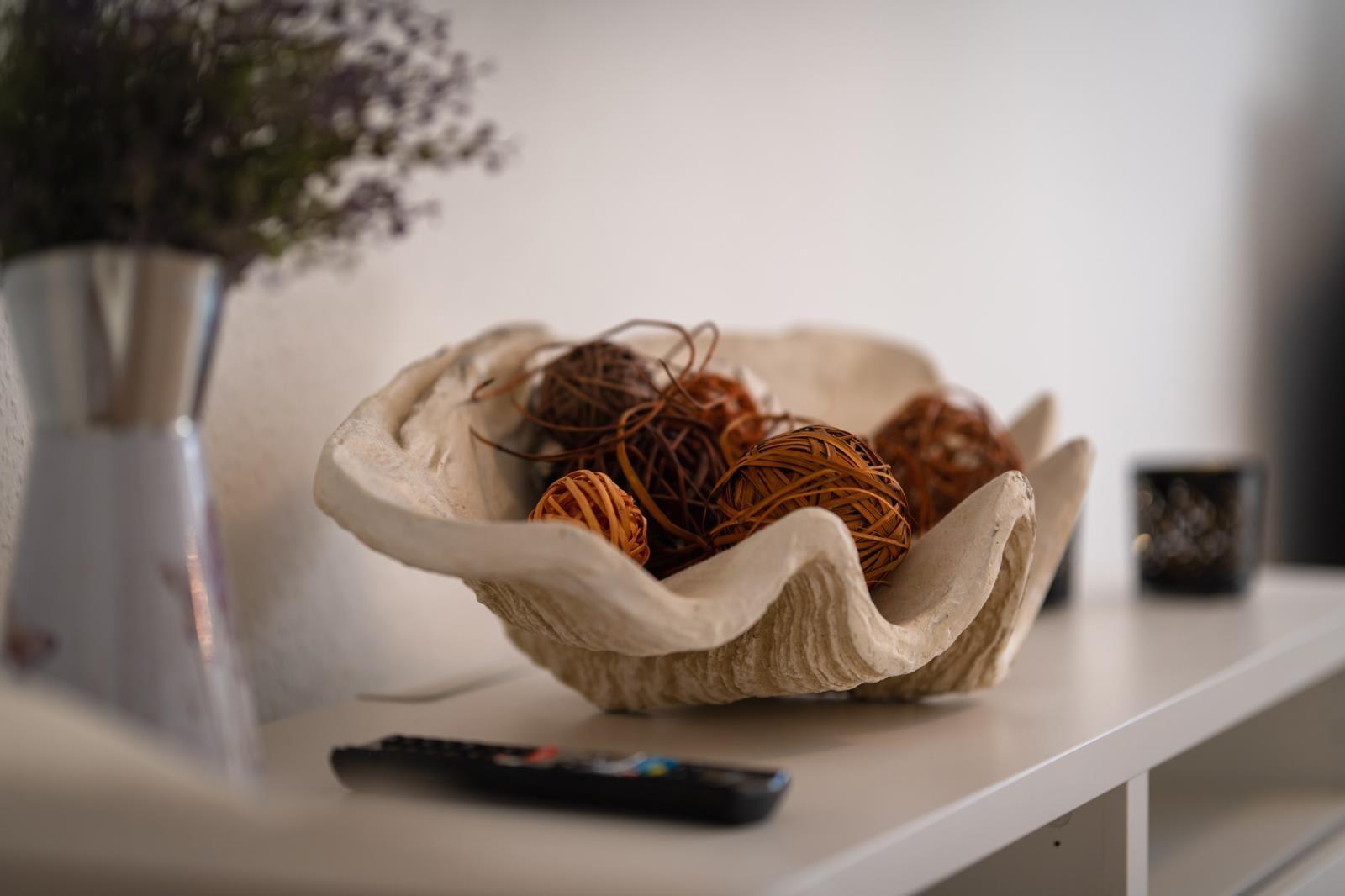 Ein Schale mit geflochtenen Kugeln steht auf einer weißen Oberfläche neben einer Vase und einem Fernbedienung.
