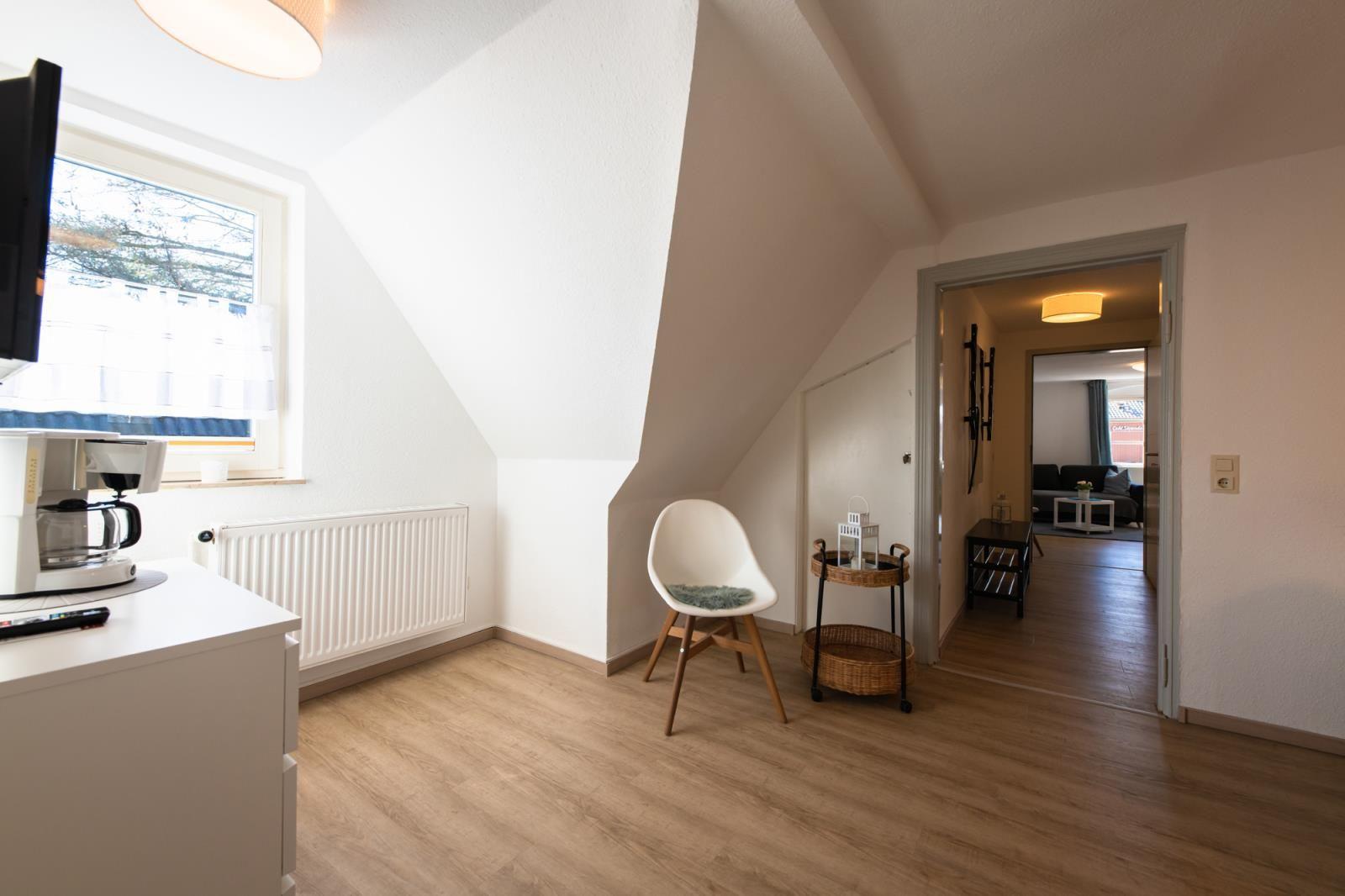 Wohnraum mit Schrägdach, Fenster, Stuhl und Blick in das Wohnzimmer.