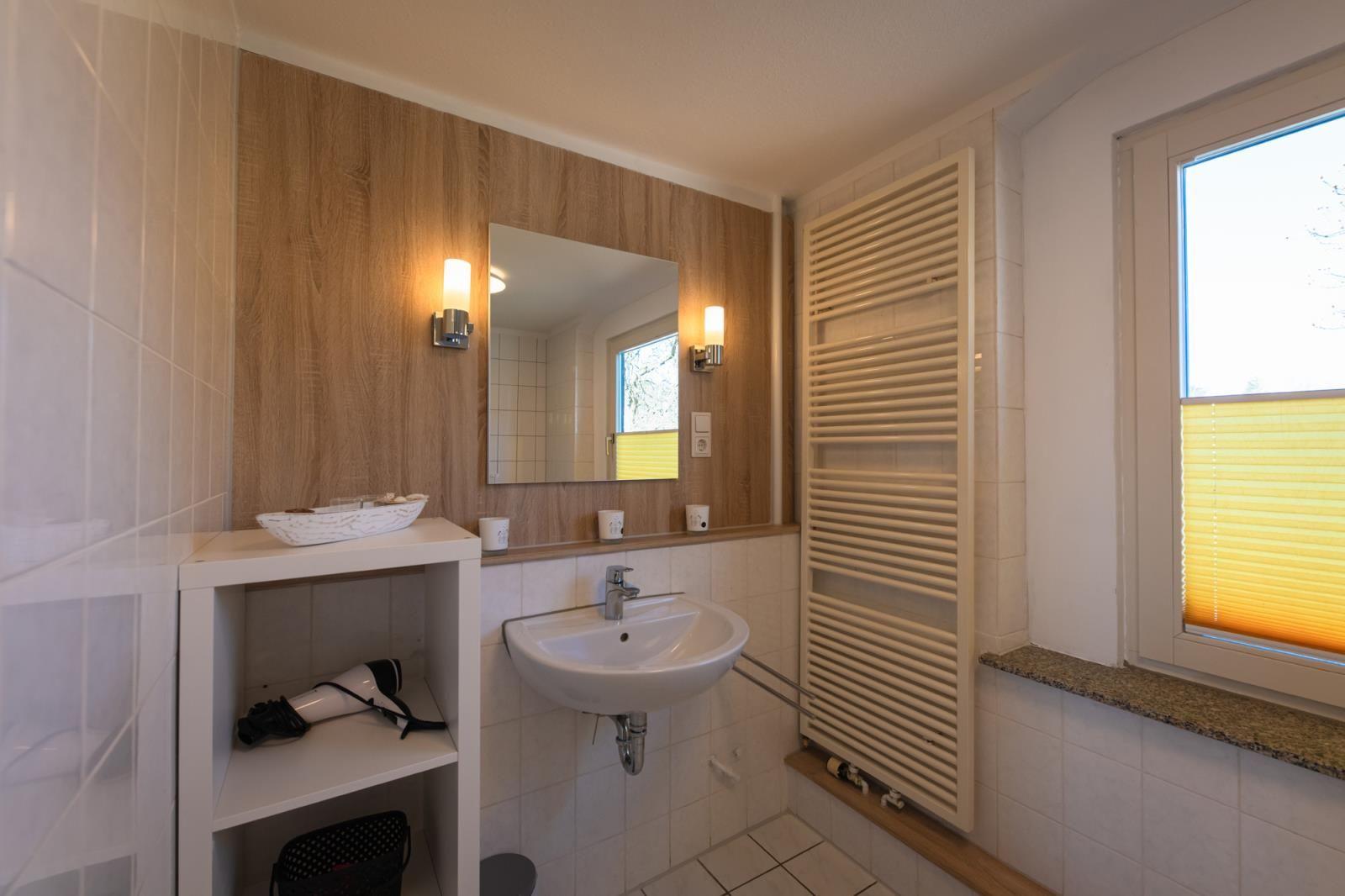 Badezimmer mit Waschbecken, Spiegel, Heizkörper und Fenster mit Jalousie.