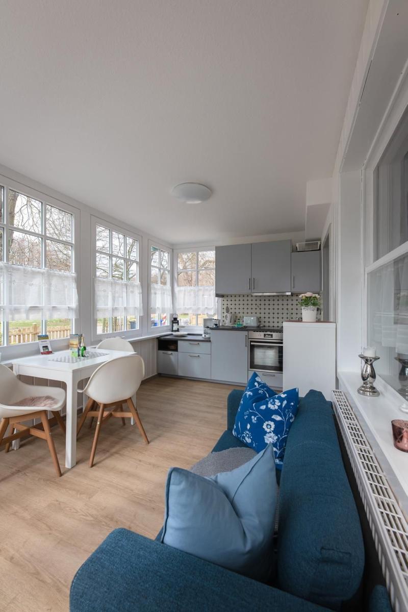 Living area with sofa, dining table, and kitchen. Large windows with white curtains.