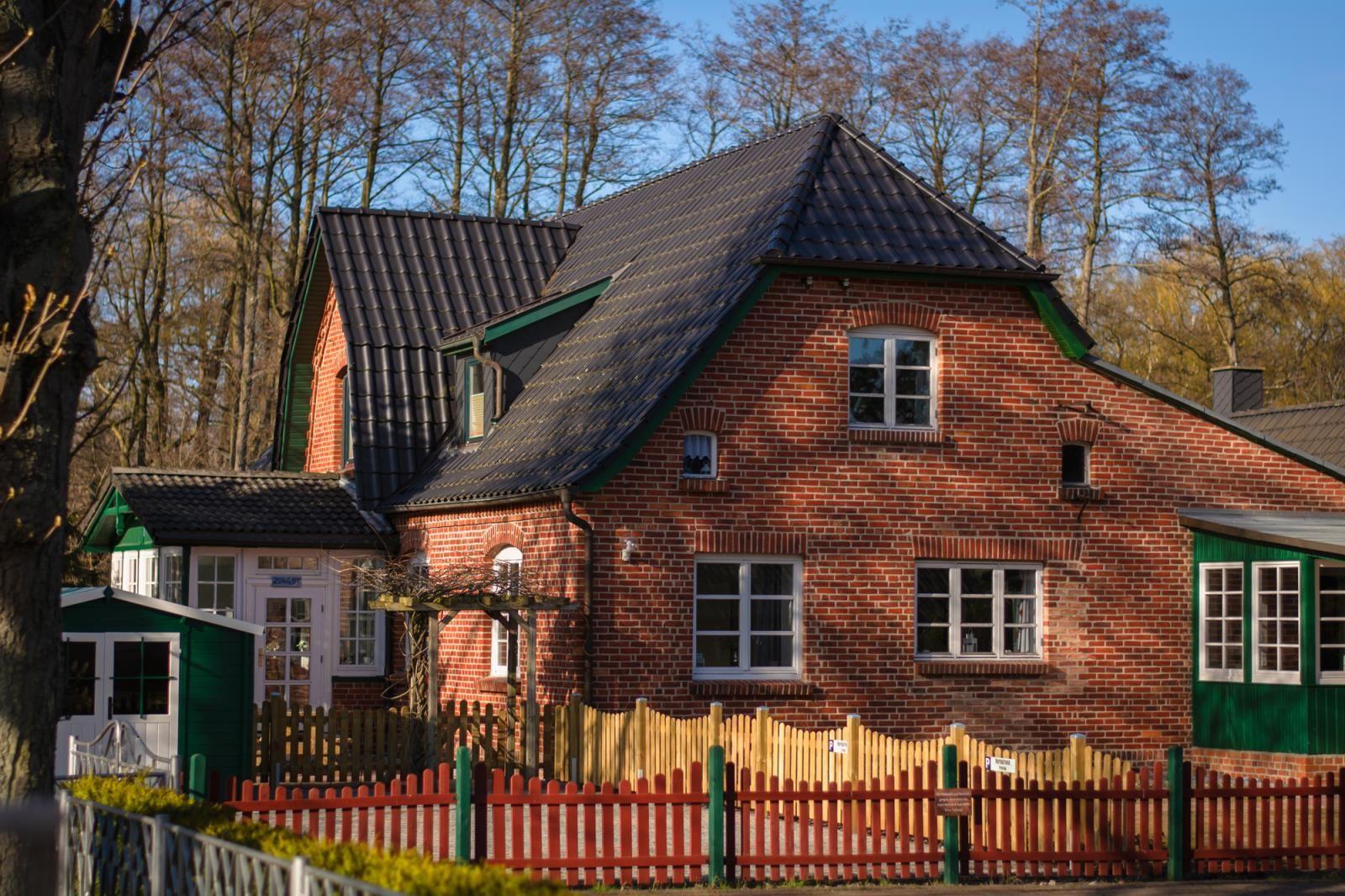 Red brick house with green accents and wooden fence. Surrounded by trees and clear sun.