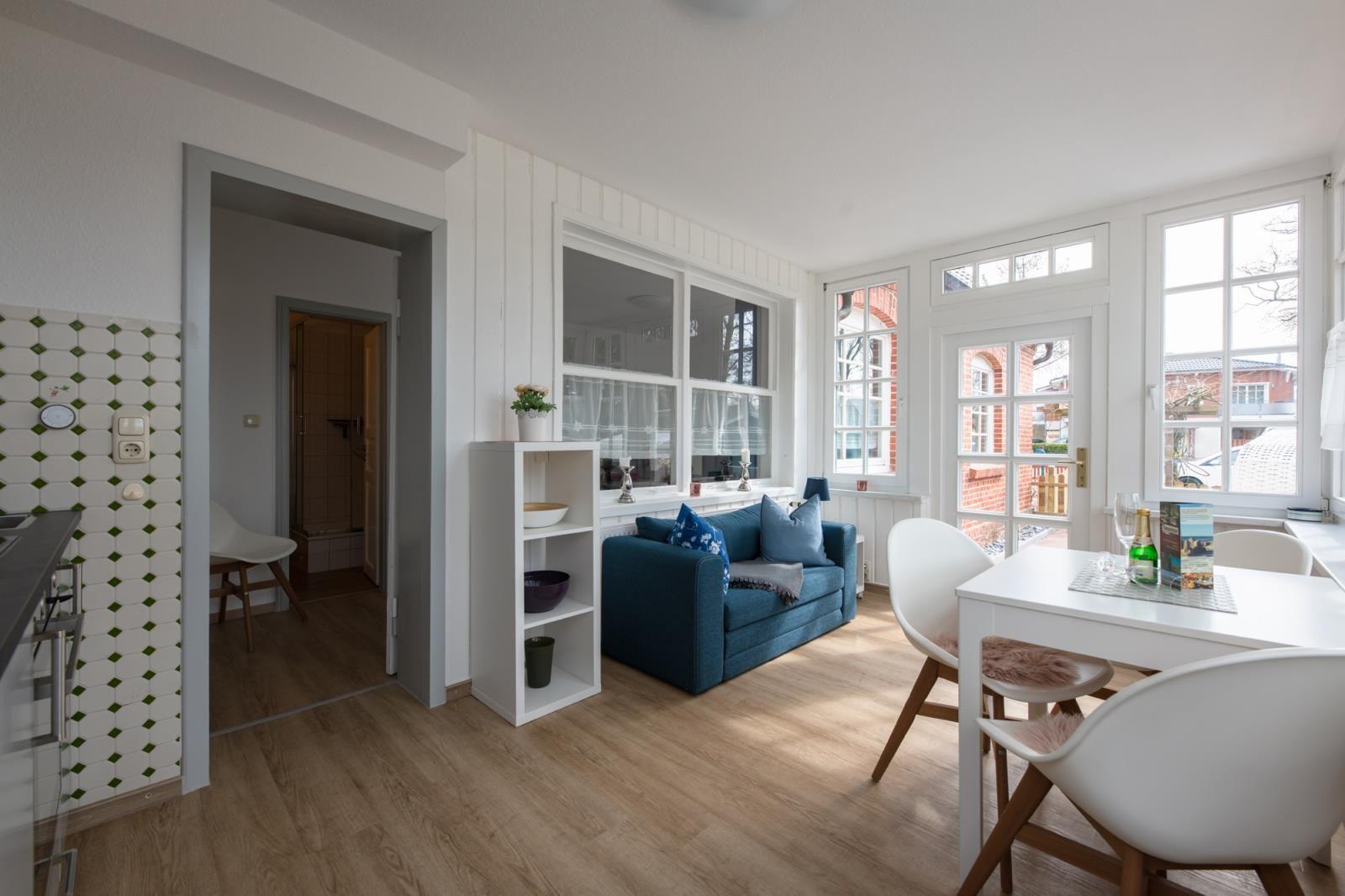 Living and dining area with blue sofa, white table and chairs, large windows and wooden floor.
