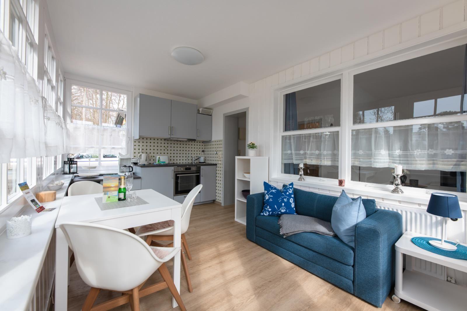 Living kitchen with blue sofa, white table, and kitchen cabinets. Large windows with white curtains.