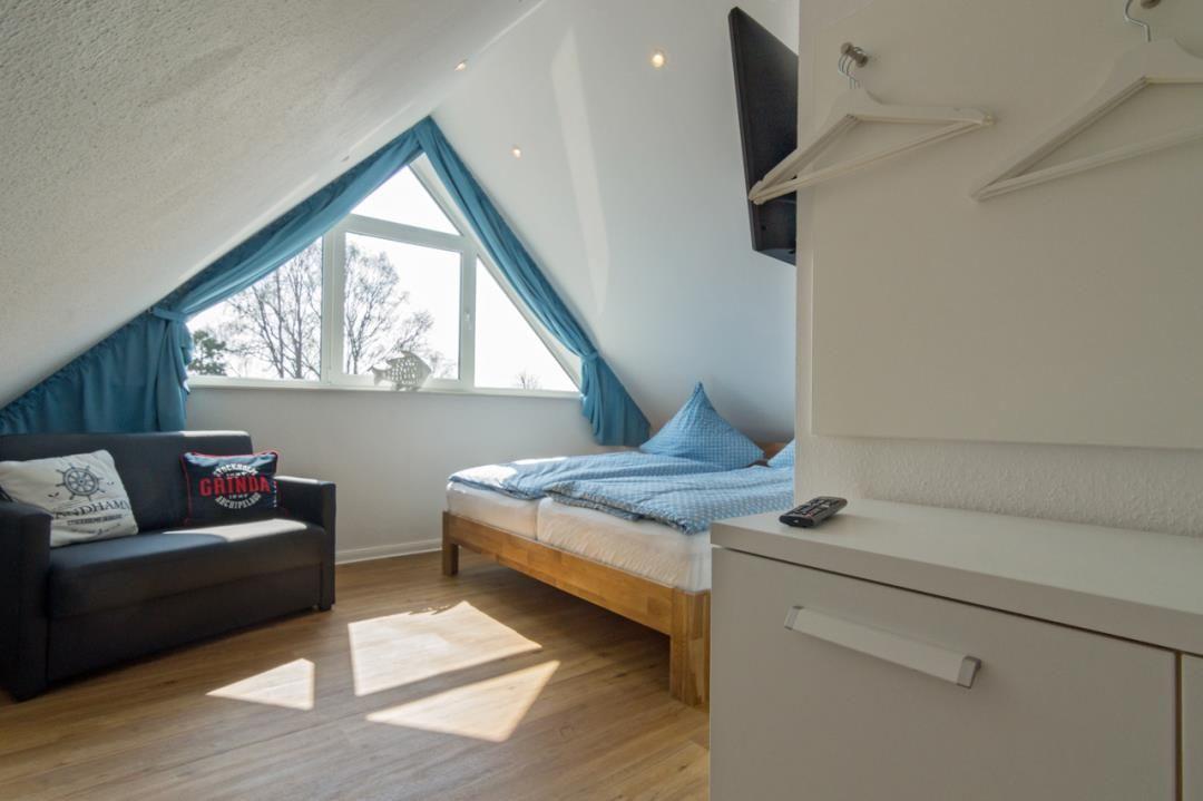 Schlafzimmer mit Dachschrägen, Bett, Sofa und TV. Fenster mit blauen Vorhängen.