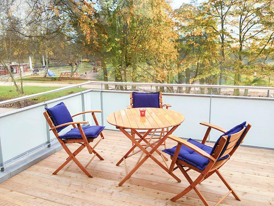 Terrasse mit Holztisch und drei Stühlen mit blauen Sitzkissen. Blick auf einen Park mit Bäumen.