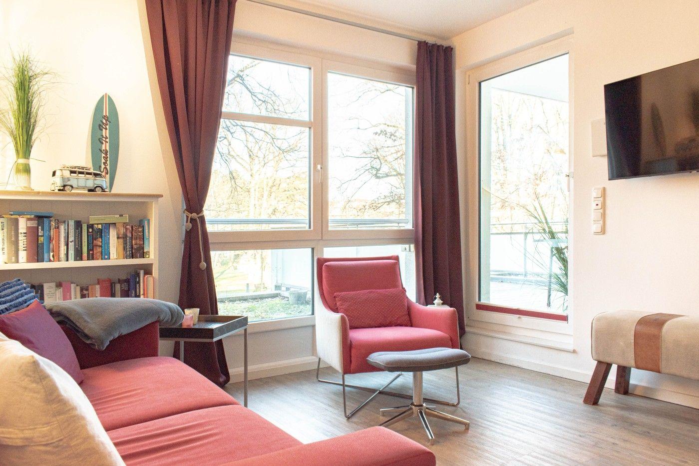 Wohnzimmer mit rotem Sofa, Sessel und großen Fenstern. Bücherregal und TV an der Wand.