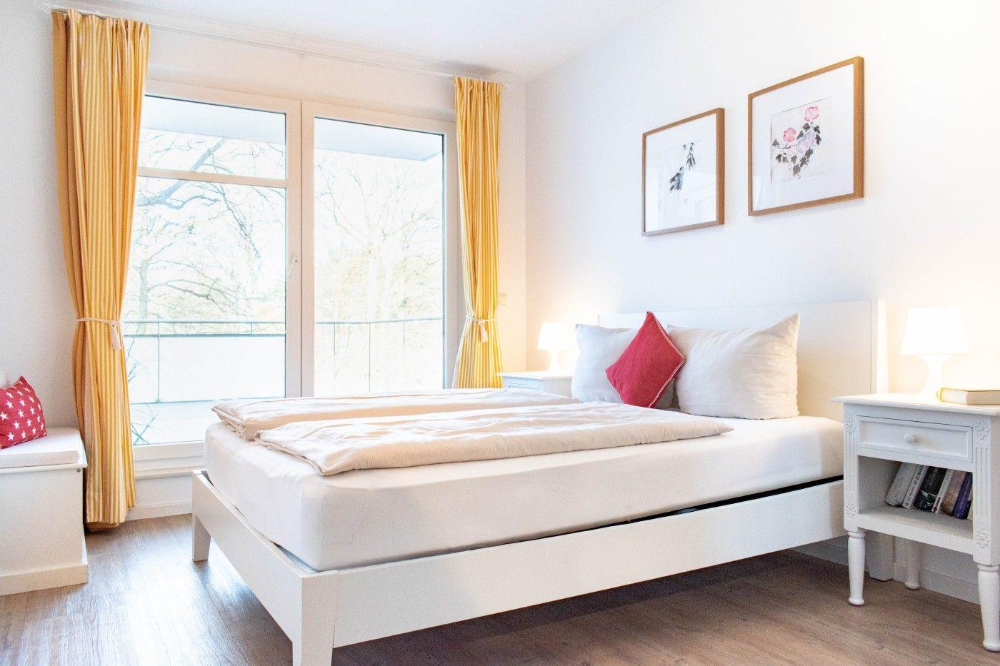 Schlafzimmer mit weißem Bett, gelben Vorhängen und Fenster zum Balkon.