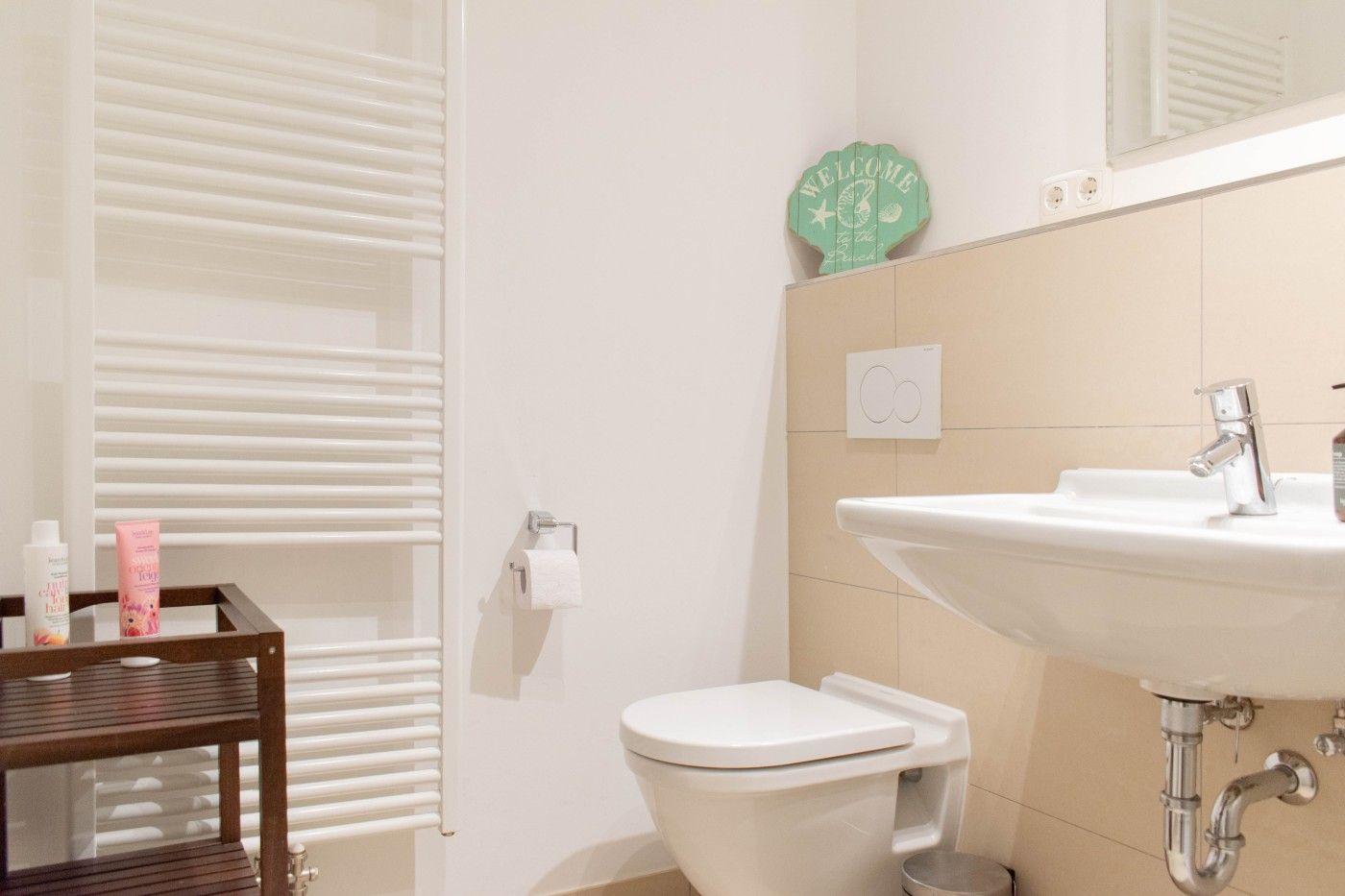 Badezimmer mit Toilette, Waschbecken und Heizkörper. Auf Wand hängt ein grünes Welcome-Schild.
