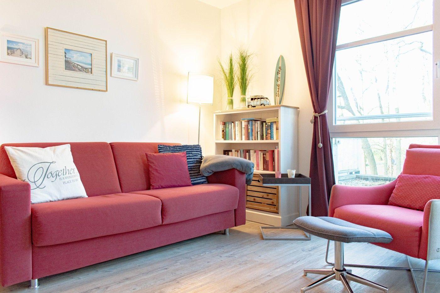 Ein Wohnzimmer mit rotem Sofa, Sessel und Bücherregal. Große Fenster mit Vorhängen.