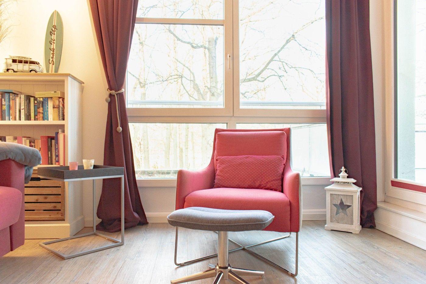 Ein Raum mit einem roten Sessel, einem Beistelltisch und einem Fenster mit Vorhängen.
