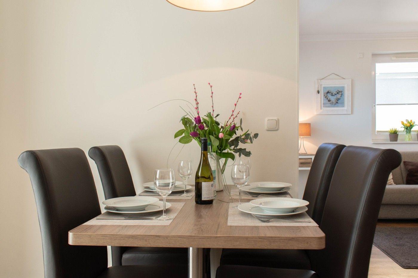 Esstisch mit vier Stühlen, Blumenvase und Weinflasche. Im Hintergrund Wohnbereich mit Sofa.