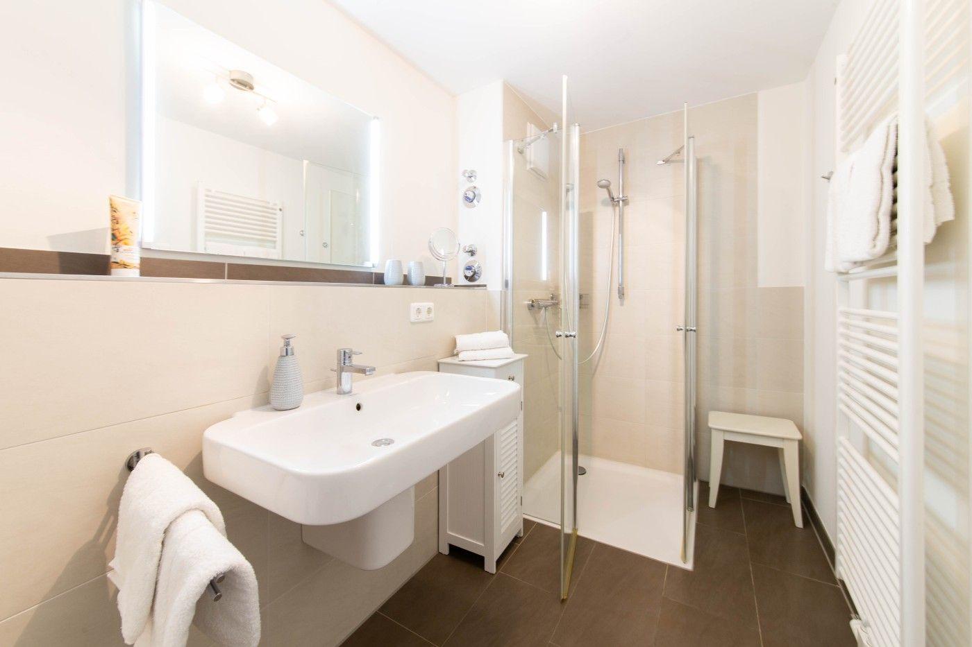 Badezimmer mit Dusche, Waschbecken und Spiegel. Handtuchhalter und Heizung sind sichtbar.