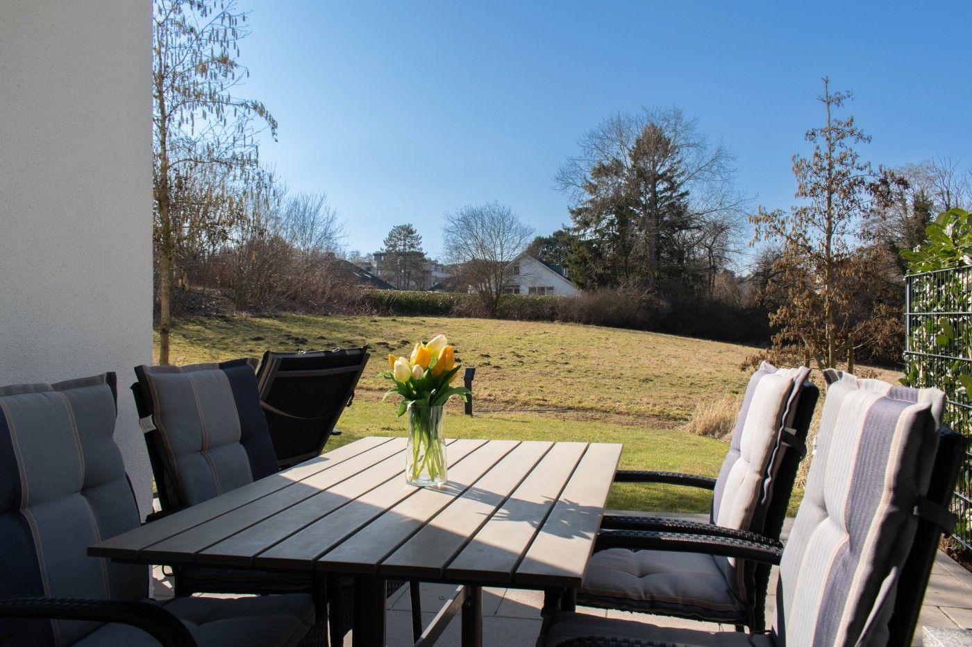 Terrasse mit Tisch und Stühlen, Blick auf grünes Feld und Bäume unter blauem Himmel.