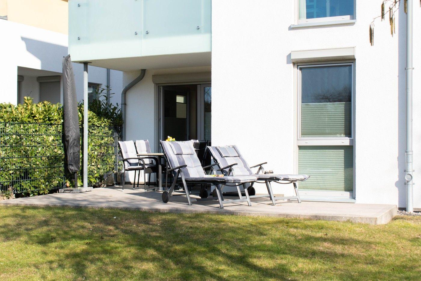 Terrasse mit Liegestühlen und Tisch vor einem weißen Haus mit Glasbrüstung.