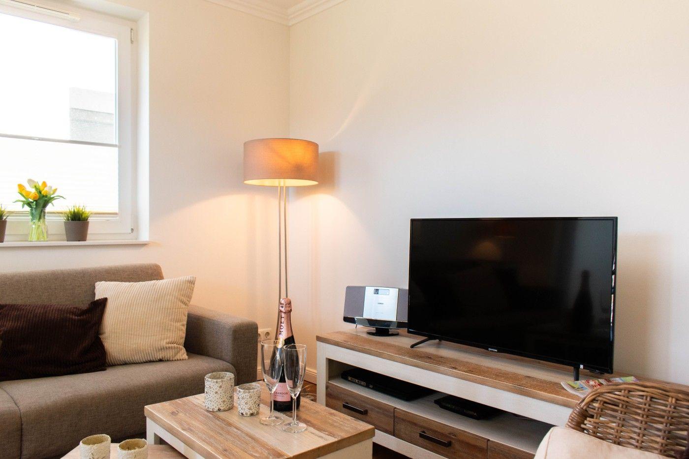 Wohnzimmer mit Sofa, Fernseher und Stehlampe. Auf dem Tisch stehen Gläser und eine Flasche.