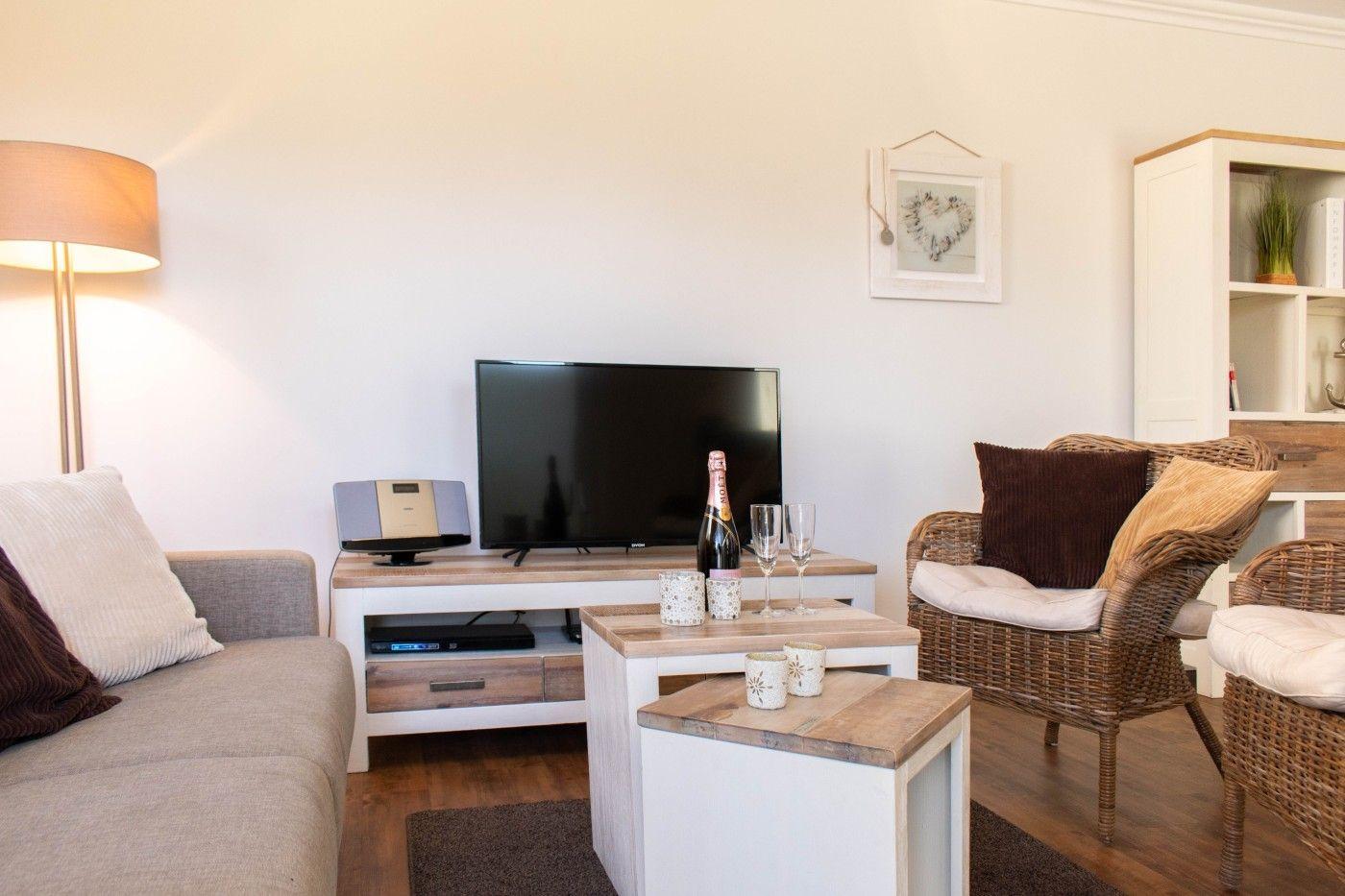 Wohnzimmer mit Sofa, Fernseher und Rattan-Stühlen. Auf dem Tisch steht eine Champagnerflasche.