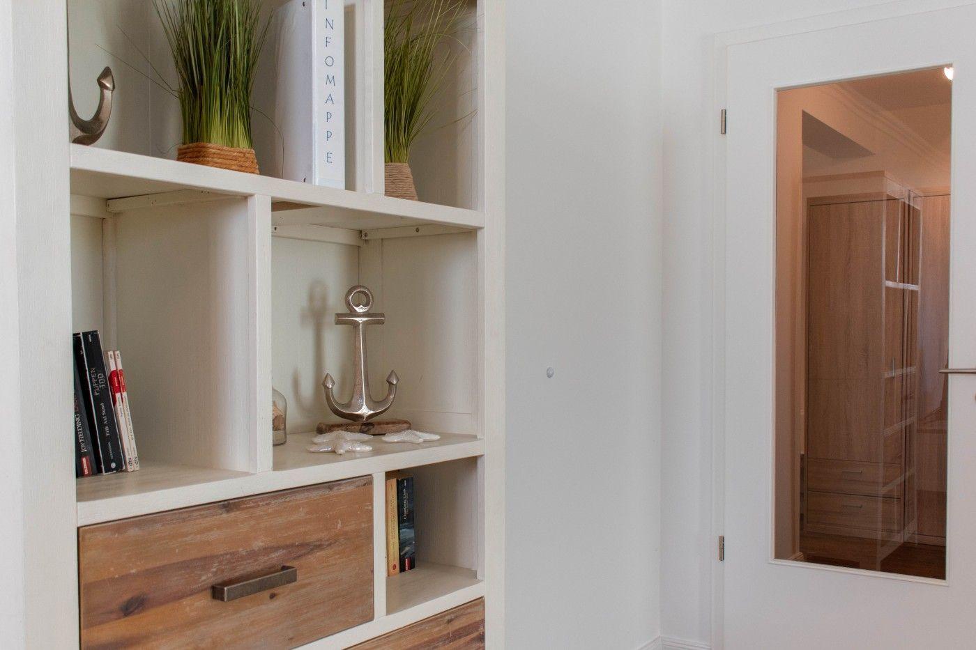 Wohnbereich mit weißen Regalen, Bücher, Pflanzen und Anker-Dekoration. Tür führt in Schlafzimmer mit Schrank.