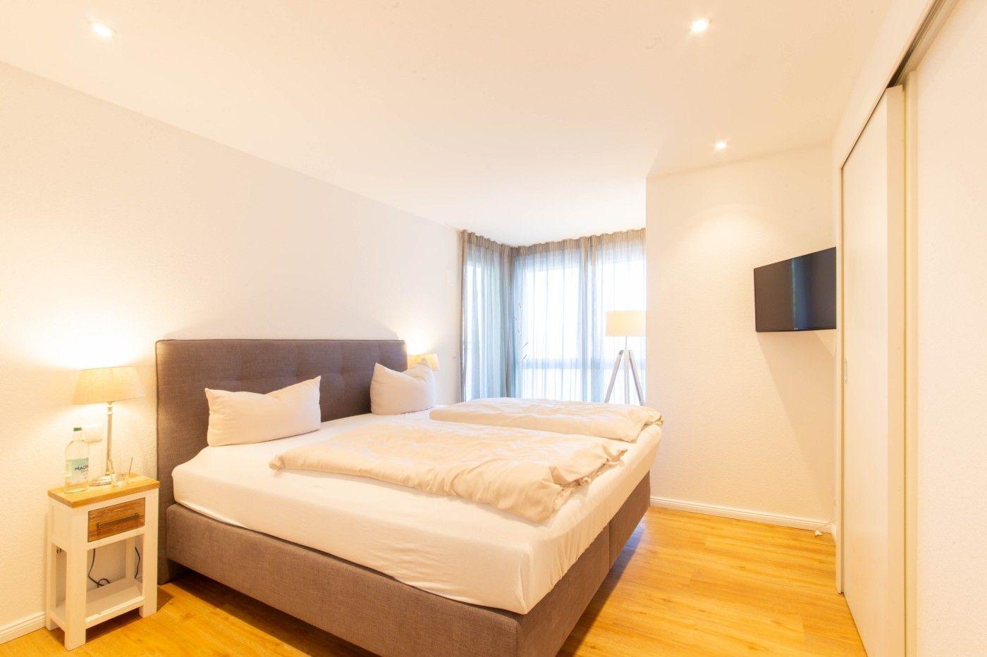 Schlafzimmer mit Doppelbett, Nachttisch, Fernseher und Fenster mit Vorhängen.