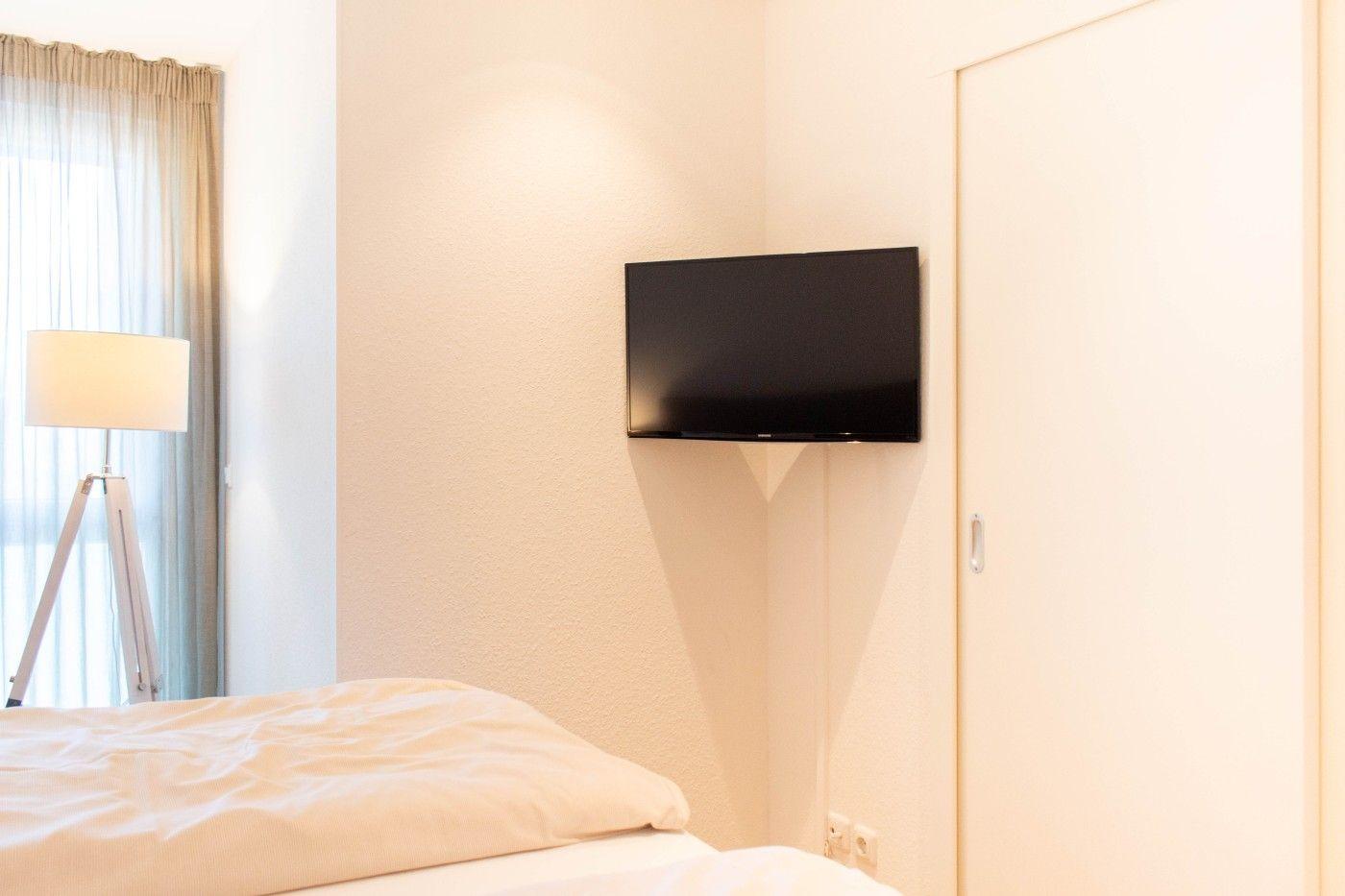 Schlafzimmer mit Bett, Wandfernseher und Stehlampe neben Fenster mit Vorhängen.