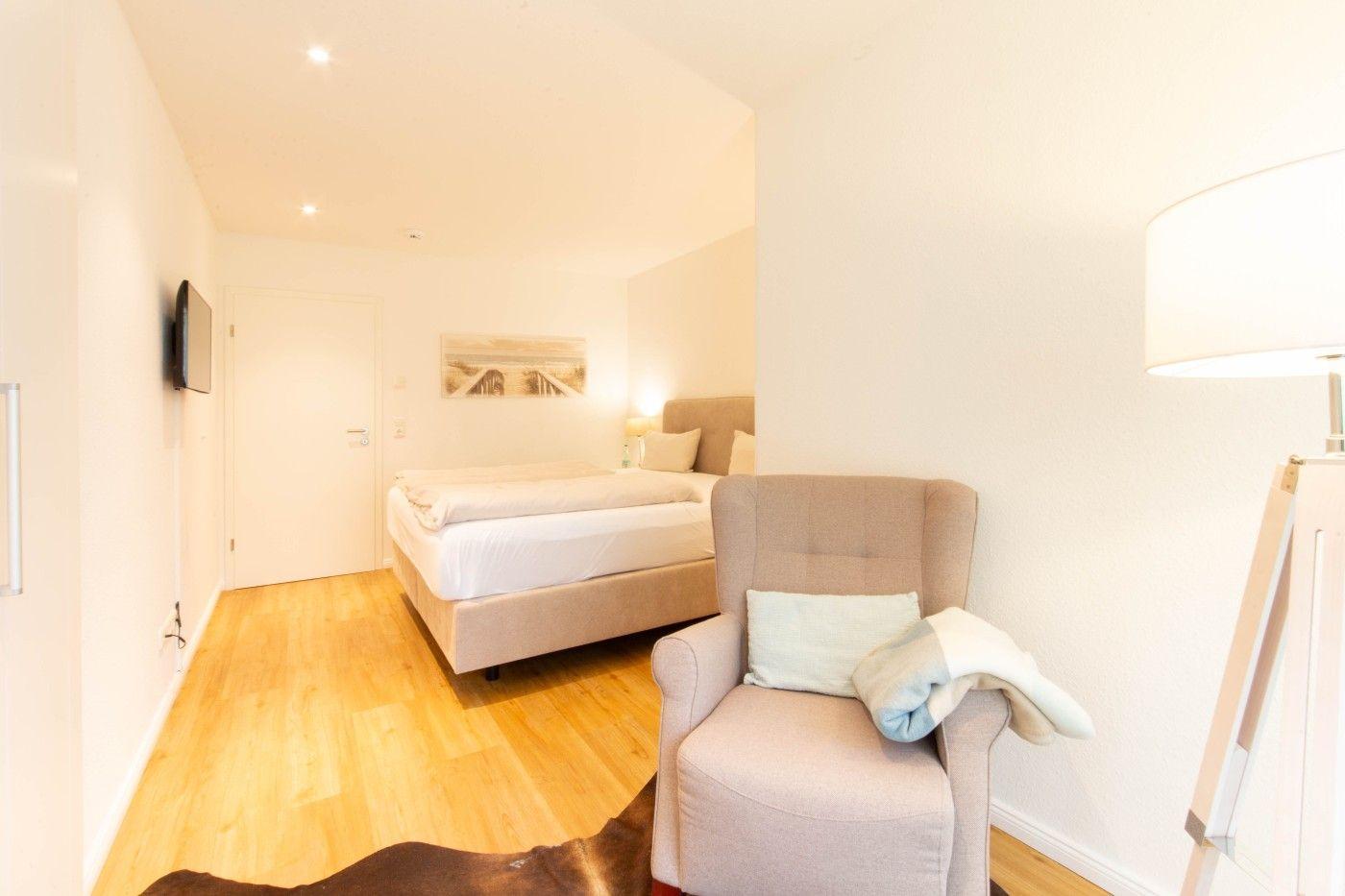 Schlafzimmer mit Bett, Sessel und Wand-TV. Holzfußboden und helle Wände.