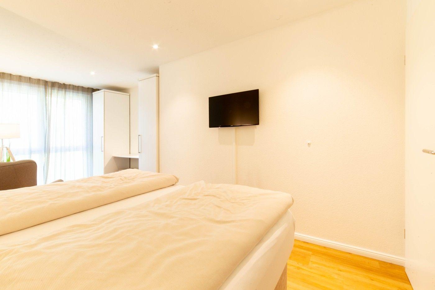 Schlafzimmer mit Bett, Wand-TV und Fenster mit Vorhängen.