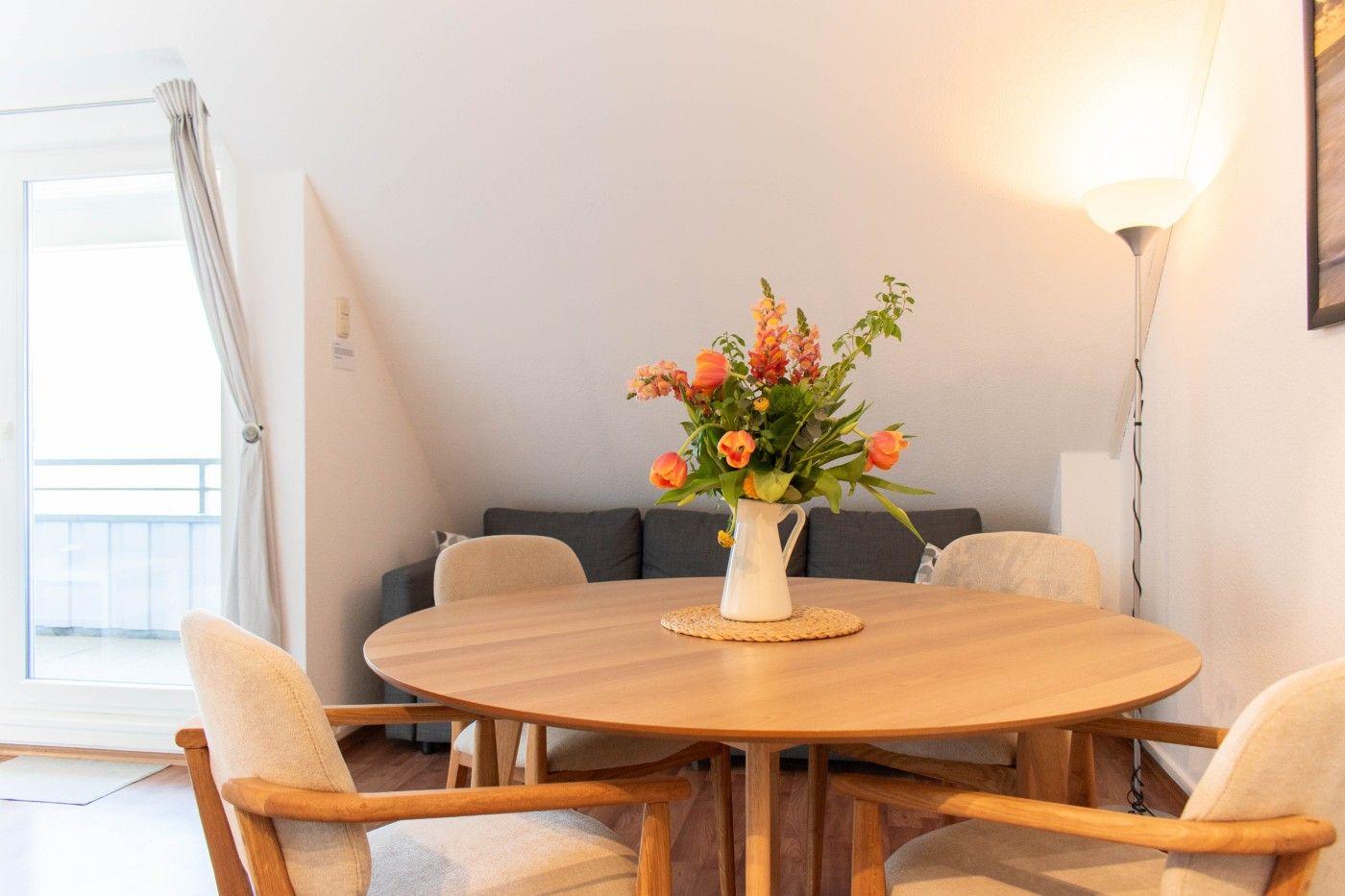 Rundes Esszimmer mit Holztisch, Stühlen und Blumenvase. Hintergrund: Sofa und Balkontür.