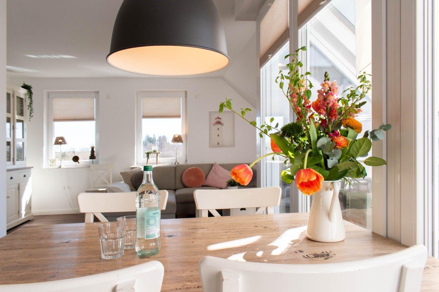 Essbereich mit Holztisch, weißen Stühlen und Blumenarrangement. Im Hintergrund Wohnzimmer mit Sofa und Fenstern.