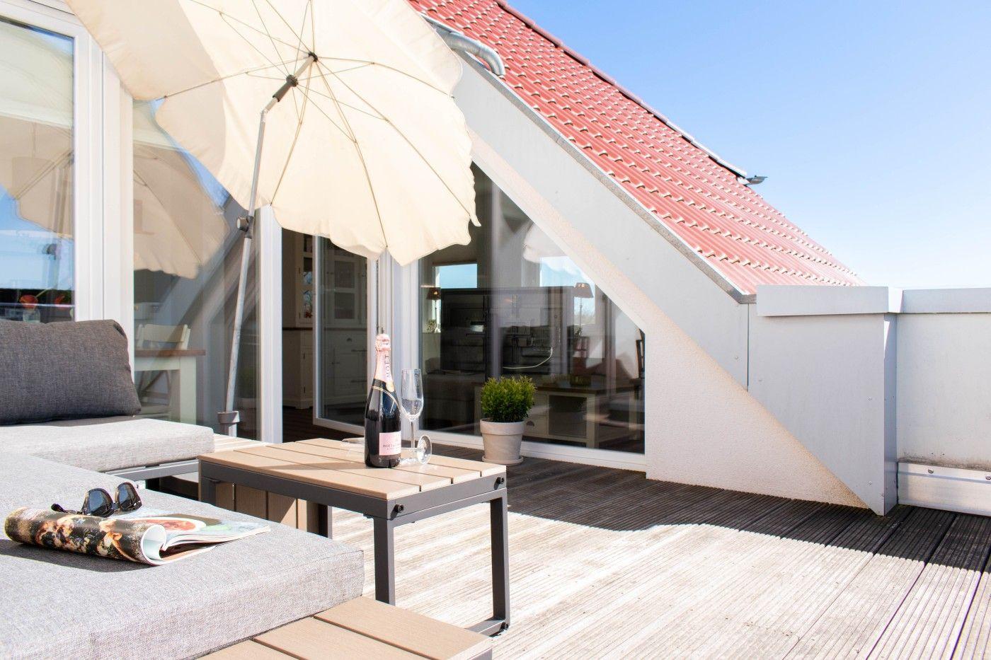 Terrasse mit Sonnenschirm, Tisch, Getränke und Sitzmöbel. Hintergrund: Haus mit rotem Dach und Glasfront.