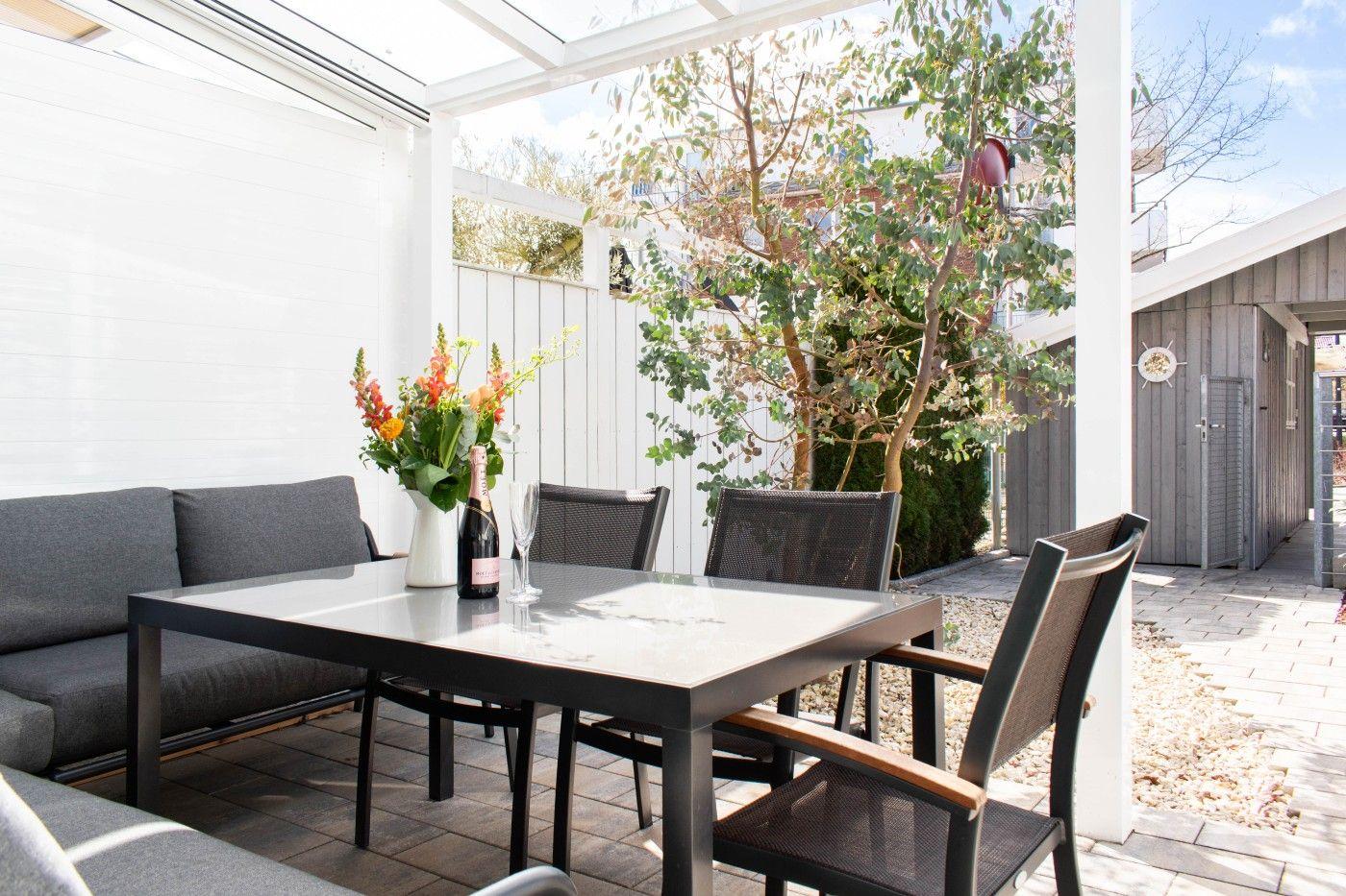 Terrasse mit Tisch, Stühlen und Sofa. Hintergrund: Baum und weißer Zaun.