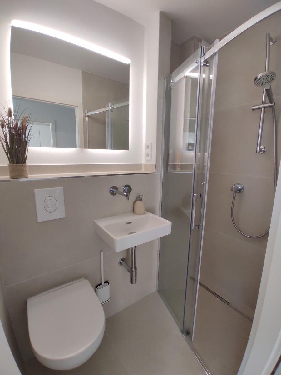 Badezimmer mit Dusche, Waschbecken und Toilette. Spiegel mit Beleuchtung.