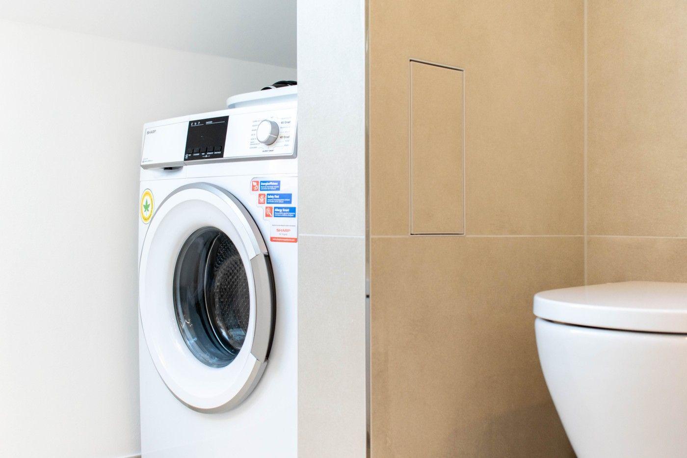Waschmaschine und Toilette in einem Raum mit hellen Wänden und beigefarbenen Fliesen.