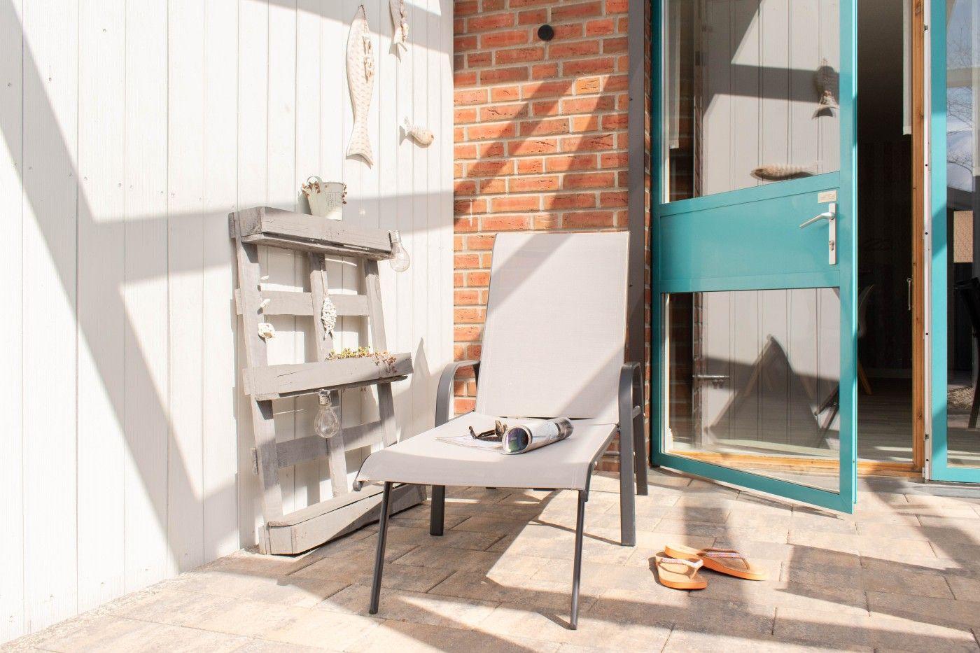Terrasse mit Liegestuhl, Holzregal und blauer Tür. Sonnenlicht fällt auf Steinplatten.